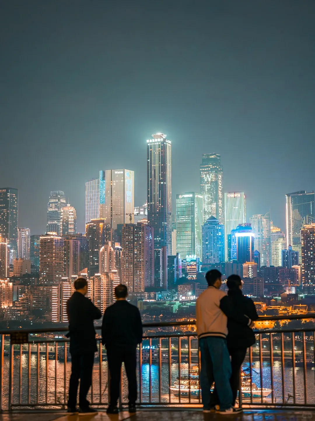 Four people standing at a railing viewing illuminated high-rise city skyline across a river at night